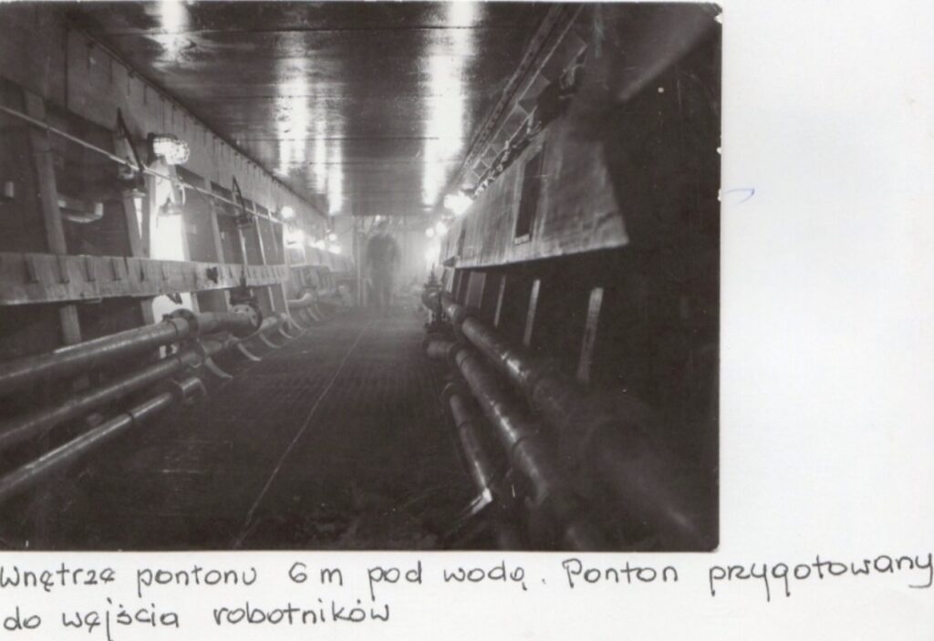 widok tunelu jaki utworzył ponton po wypompowaniu wody balastowej z jego zbiorników balastowych i z przestrzeni roboczej, skutkiem czego ponton został dociśnięty do obu połówek kadłuba, tworząc szczelne połączenie. Po prawej widoczny kanał wentylacyjny dostarczający świeże powietrze dla pracujących tam najpierw spawaczy, potem zaś malarzy. Biegnące rury przy ścianach to rury systemu balastowego pontonu.